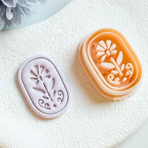 May include: Two floral-shaped cookie cutters. One is a light lilac, the other is a peach colour. Both are oval-shaped with a flower and leaf design. The cutters are on a white surface.
