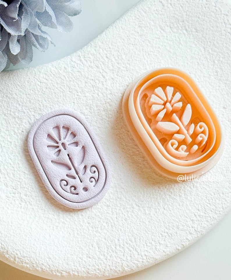 May include: Two floral-shaped cookie cutters. One is a light lilac, the other is a peach colour. Both are oval-shaped with a flower and leaf design. The cutters are on a white surface.