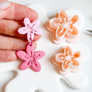 May include: Three pink and peach flower-shaped cookie cutters. The cutters are on a white surface. The largest cutter is peach, the medium cutter is pink, and the smallest cutter is a darker pink. The cutters are for making cookies or fondant decorations. The text "@lulicutters" is visible in the bottom right corner of the image.