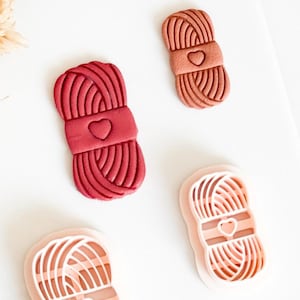 Op de afbeelding: Vier koekjessnijders met een garen-thema in tinten rood en perzik. Elke snijder heeft een hartvormig detail op de band rond het garen. De snijders zijn ontworpen om koekjes in de vorm van bollen garen te maken.