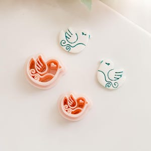 Op de afbeelding: Vier vogelvormige items. Twee zijn lichtroze koekjessnijders en twee zijn wit met teal details. De koekjessnijders hebben een gedetailleerd vogelontwerp. De andere twee vogels hebben teal vleugel- en staartdetails.