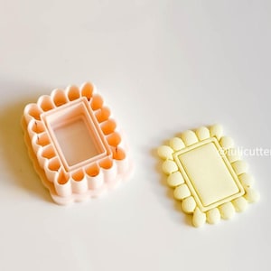 May include: Two cookie cutters, one pink and one yellow, each in a rectangular shape with a scalloped border. The pink cutter is stacked, showing the depth of the design. The yellow cutter is a finished cookie.