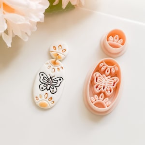 Może przedstawiać: Białe i pomarańczowe kolczyki z gliny polimerowej z motywem motyla i pasującymi wykrawaczkami do gliny. Kolczyki mają złoty drucik i czarny motyw motyla. Wykrawaczki są jasnoróżowe.