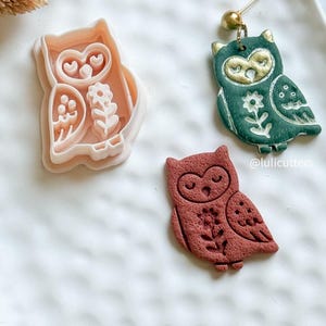 May include: An assortment of owl-themed items. A light pink owl-shaped cookie cutter, a dark green and gold owl earring, and a reddish-brown owl-shaped clay piece. All items feature floral and heart details.