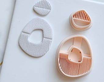 May include: A collection of polymer clay cutters in a neutral color palette. The cutters are in various organic shapes, including teardrops and rounded forms, with textured and ribbed details. The shapes are designed for crafting earrings or other jewelry.