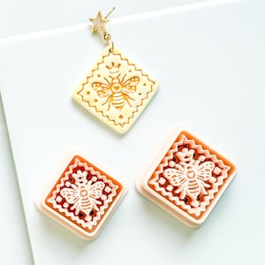 May include: A set of bee-themed jewelry and craft supplies. The image features a gold star earring, a cream-colored pendant with a bee design, and two square stamps with bee motifs. The bee designs are detailed and the items are set against a white background.