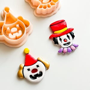 Puede incluir: Una colección de artículos con temática de payasos. Incluye dos cortadores de galletas de plástico rosa con forma de cabeza de payaso y una cara de payaso con sombrero. También, una cara de payaso pintada con sombrero rojo y cuello morado.