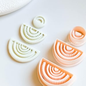 May include: A collection of white and peach-colored cookie cutters in a variety of shapes. The cutters are arranged on a white surface. The shapes include a half-circle with interior details and a small circle.