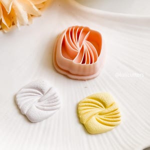 Pode incluir: Um cortador de biscoitos de plástico rosa com um design em espiral está ao lado de dois biscoitos, um branco e um amarelo, também com designs em espiral. O cortador de biscoitos e os biscoitos estão em uma superfície branca.