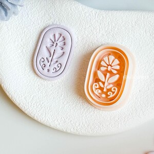 May include: Two floral-shaped cookie cutters. One is light purple, the other orange. Both are oval with a flower design. The cutters are on a white, textured surface.