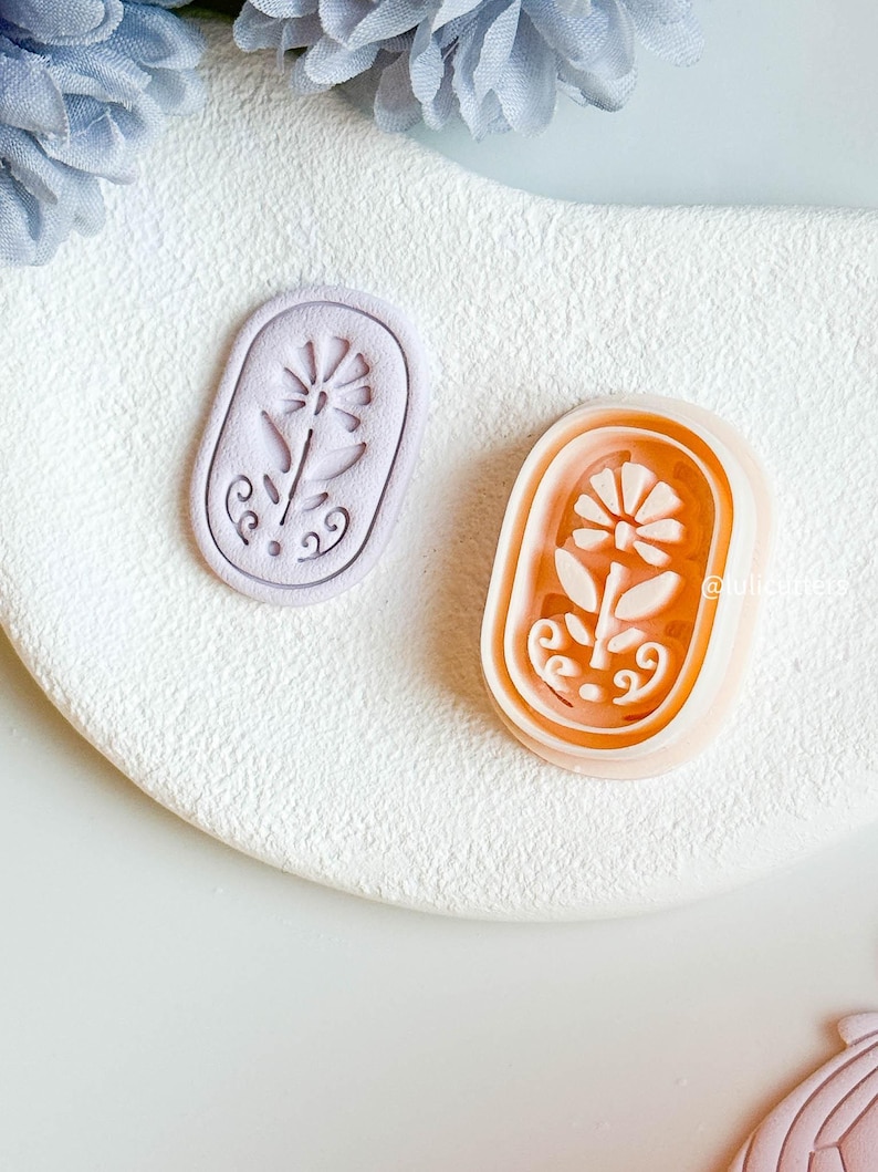 May include: Two floral-shaped cookie cutters. One is light purple, the other orange. Both are oval with a flower design. The cutters are on a white, textured surface.