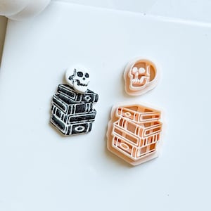 Könnte beinhalten: Eine Sammlung von Halloween-Ausstechern. Ein Ausstecher zeigt einen Bücherstapel mit einem Totenkopf, ein weiterer einen Totenkopf und der dritte einen Bücherstapel. Die Ausstecher sind in Schwarz, Weiß und Pfirsich gehalten.