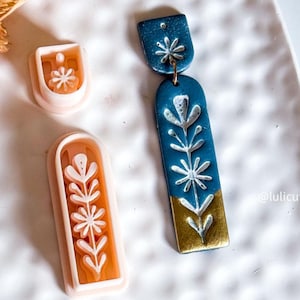Könnte beinhalten: Ein Paar blau-goldene Ohrringe mit floralem Design und zwei Ton-Ausstechern. Die Ohrringe haben eine rechteckige Form mit weißen Blumenmustern. Die Ausstecher sind in Form der Ohrringe und einer Blume.