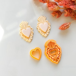 Può includere: Una collezione di formine per biscotti a forma di cuore di varie dimensioni. Le formine sono in tonalità di giallo e arancione, con un design a cuore rosa chiaro. L'immagine include anche due biscotti a forma di cuore.