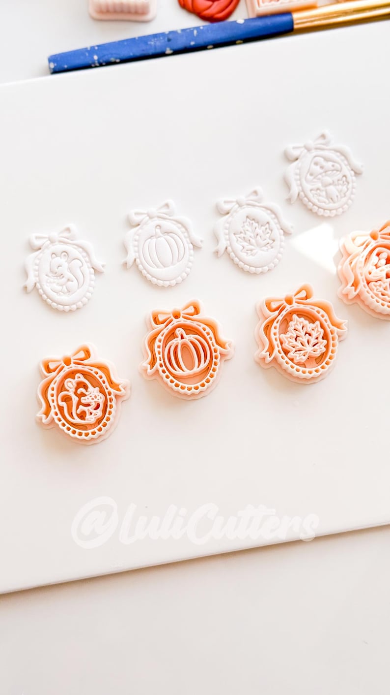 May include: A collection of oval-shaped cookie cutters featuring embossed designs of a squirrel, a pumpkin, a maple leaf, and a mushroom, each topped with a bow. The cutters are white and peach-colored, with the text @LuliCutters visible at the bottom.