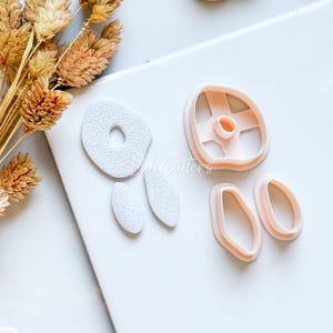 May include: A collection of light pink cookie cutters in various shapes, including a circle, oval, and a flower. The cutters are arranged on a white surface, with dried wheat stalks in the upper left corner. The image is well-lit.