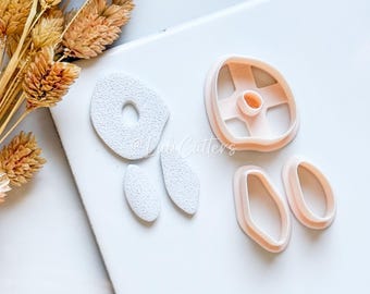 May include: A collection of light pink cookie cutters in various shapes, including a circle, oval, and a flower. The cutters are arranged on a white surface, with dried wheat stalks in the upper left corner. The image is well-lit.