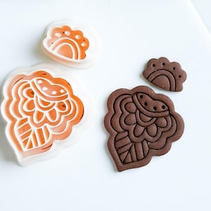 Puede incluir: Vista aérea de tres cortadores de galletas y una galleta. Los cortadores son blancos con interiores naranjas, y la galleta es marrón. Los diseños son florales y geométricos. Flores secas en el fondo.