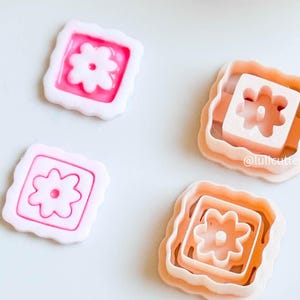 Könnte beinhalten: Vier blütenförmige Ausstecher. Zwei sind rosa und weiß, mit einem Blumendesign in einem quadratischen Rahmen. Die anderen beiden sind pfirsichfarben, ineinander verschachtelt und weisen ebenfalls ein Blumendesign auf.