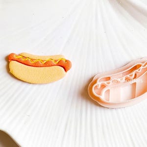 May include: A close-up of two hot dog cookie cutters. One is a light orange color and the other is a light pink color. The cutters are on a white plate with a textured surface.