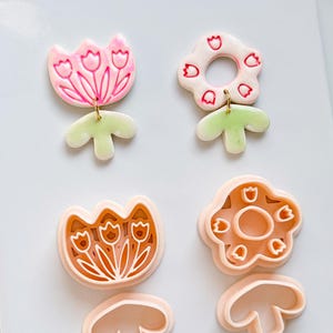 May include: Floral-themed earrings and matching cookie cutters. The earrings feature pink and white flower designs with gold-coloured hooks and green leaf accents. The cookie cutters are in the shape of flowers and leaves.