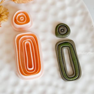 Può includere: Due taglierini in argilla polimerica a forma di orecchini astratti. Uno è color pesca e arancione, l'altro verde oliva. I taglierini hanno un design a strati, con il testo "@lulicutters" visibile sul lato destro.