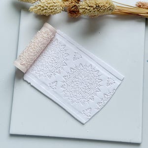 Può includere: Una lastra di argilla bianca con un motivo floreale in rilievo e un rullo rosa chiaro con un motivo abbinato. Spighe di grano essiccate sono disposte su una superficie bianca, creando una composizione incentrata sull'artigianato.