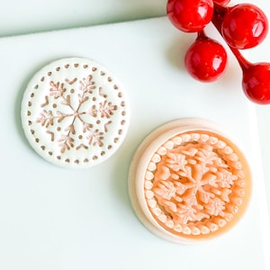 May include: Two circular decorative items with snowflake designs. One is white with a brown snowflake and dotted edge. The other is light brown with a raised snowflake pattern. Red berries are in the background.