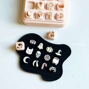 May include: A collection of Halloween-themed cookie cutters in a light pink container and on a black surface. The cutters include shapes like pumpkins, skulls, bats, spiders, and witch hats. The image is taken from above.