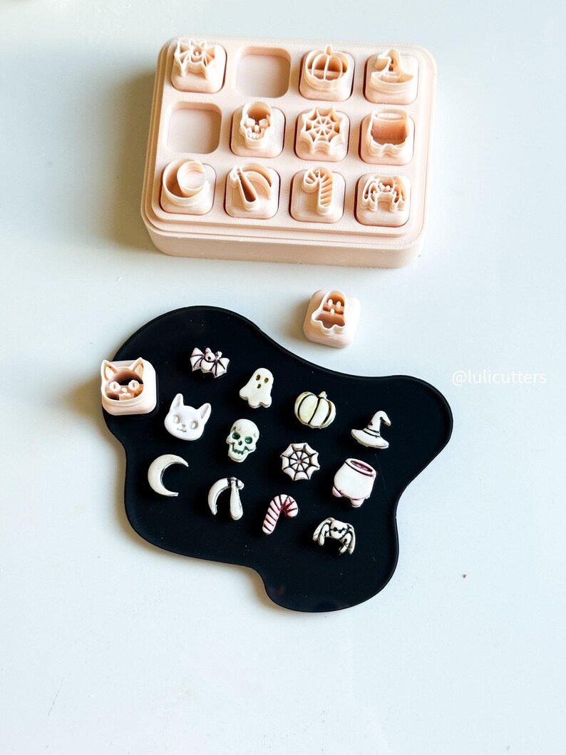 May include: A collection of Halloween-themed cookie cutters in a light pink container and on a black surface. The cutters include shapes like pumpkins, skulls, bats, spiders, and witch hats. The image is taken from above.