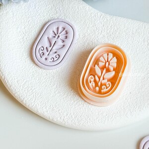 May include: Two floral-shaped cookie cutters. One is a light purple colour, and the other is a peach colour. Both are oval-shaped with a flower design in the centre. The cutters are on a white, textured surface.