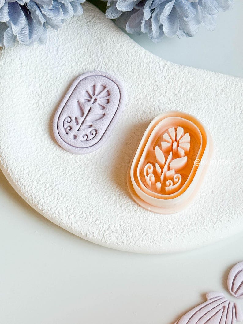 May include: Two floral-shaped cookie cutters. One is a light purple colour, and the other is a peach colour. Both are oval-shaped with a flower design in the centre. The cutters are on a white, textured surface.