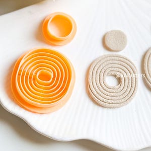 May include: A set of four round cookie cutters, two are beige and two are orange. The orange cutters are concentric circles, while the beige cutters are solid. The cutters are on a white plate with a wavy edge.