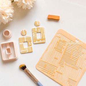 May include: An overhead shot displays crafting tools and supplies. The image features a light blue handled brush, a stencil, and several polymer clay earring components in shades of white and yellow. The @lulicutters watermark is visible on the stencil.