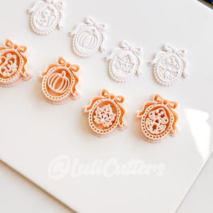 May include: A collection of white and peach-colored plastic cookie cutters shaped like oval frames with bows. The designs include a squirrel, a pumpkin, a maple leaf, and a mushroom. The text @LuliCutters is visible at the bottom of the frame.