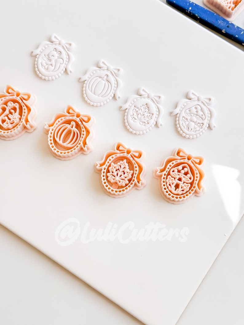 May include: A collection of white and peach-colored plastic cookie cutters shaped like oval frames with bows. The designs include a squirrel, a pumpkin, a maple leaf, and a mushroom. The text @LuliCutters is visible at the bottom of the frame.