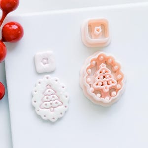 May include: A collection of pink and white Christmas tree-shaped cookie cutters and a finished cookie. The cutters are in various shapes, including a square and a scalloped circle. The finished cookie is white with a pink tree design.