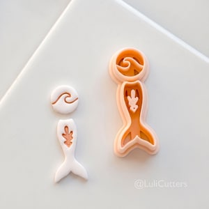 May include: Two cookie cutters and a cookie, one in the shape of a mermaid tail and the other in the shape of a wave. The cookie cutters are peach-colored, and the cookie is white. The image includes the text "@LuliCutters".