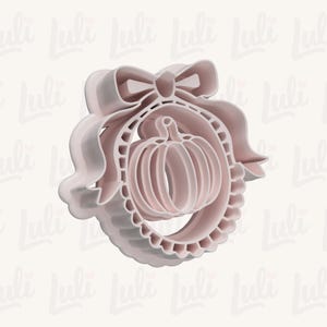 May include: A light pink plastic cookie cutter featuring a central pumpkin shape enclosed within a circular frame that is decorated with a ruffled edge and topped with a large, prominent bow detail at the top of the circular structure.