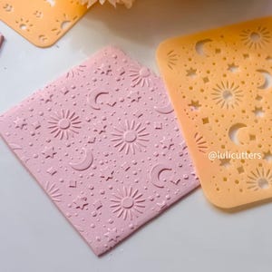 May include: Close-up of pink and orange stencils with celestial designs. The pink stencil has embossed suns, moons, and stars. The orange stencil has cut-out suns, moons, and stars. The text "@lulicutters" is visible on the orange stencil.