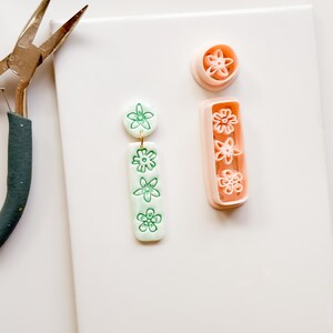 May include: A white surface displays crafting tools and materials. A pair of pliers, a gold earring, and two floral-shaped clay cutters are visible. A pair of green and white floral earrings are also present, showcasing a handmade aesthetic.