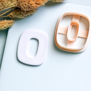 Könnte beinhalten: Ein weißer, strukturierter Buchstabe "O" und ein pfirsichfarbener Ausstecher in Form des Buchstabens "O". Der Ausstecher hat eine quadratische Außenform mit abgerundeten Ecken. Getrocknete Weizenähren sind im Hintergrund.