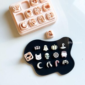May include: A collection of Halloween-themed cookie cutters in a light pink container and on a black surface. The cutters include pumpkins, ghosts, bats, spiders, and other spooky shapes. The image is taken from above.