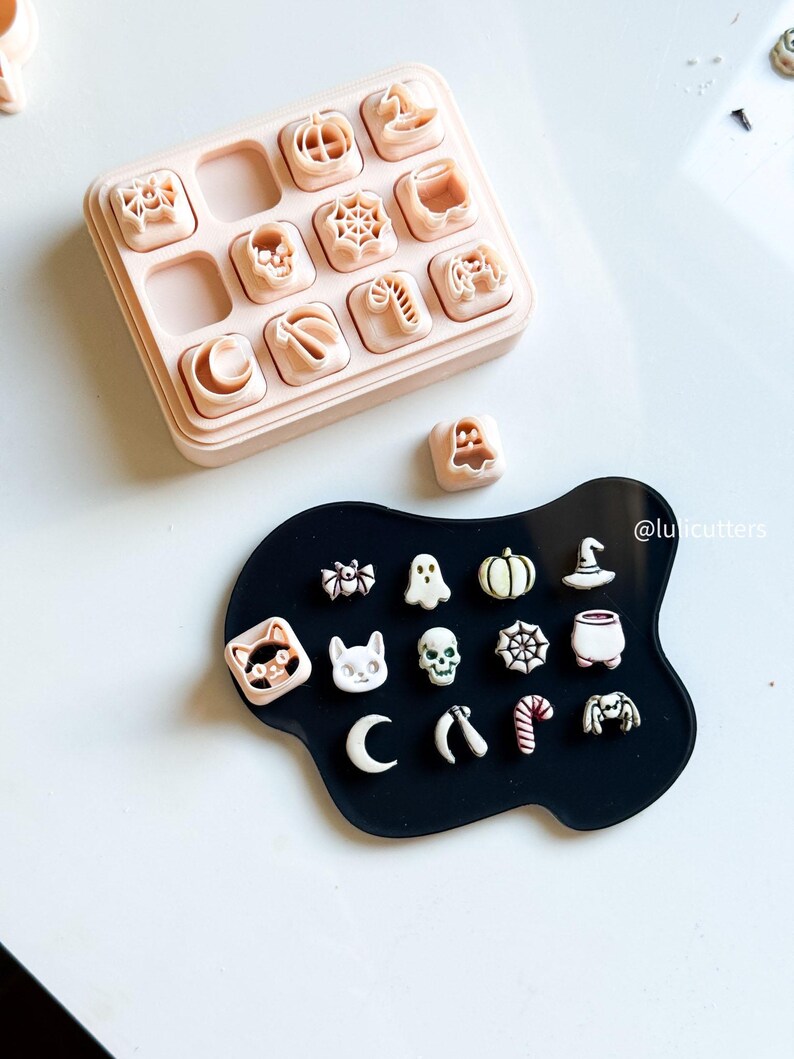 May include: A collection of Halloween-themed cookie cutters in a light pink container and on a black surface. The cutters include pumpkins, ghosts, bats, spiders, and other spooky shapes. The image is taken from above.