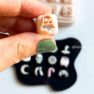 May include: A small, beige ghost-shaped cookie cutter held in a hand, with a tray of other cutters in the background. The cutter is held over a black surface with other small cutters. The text "@lulicutters" is visible.