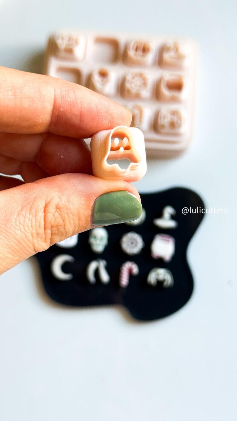May include: A small, beige ghost-shaped cookie cutter held in a hand, with a tray of other cutters in the background. The cutter is held over a black surface with other small cutters. The text "@lulicutters" is visible.