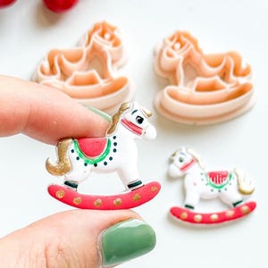 Pode incluir: Miniaturas de enfeites de cavalo de balanço. Uma é segurada entre os dedos, mostrando um cavalo branco com sela e base vermelhas, detalhes dourados e detalhes verdes e pretos. Outros enfeites são de cor pêssego.