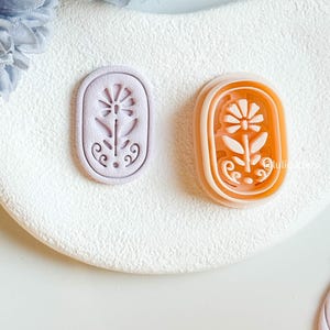 May include: Two floral-shaped cookie cutters. One is lavender, the other is orange with a white interior. Both are oval-shaped with a flower and stem design. The cutters are on a white, textured surface.
