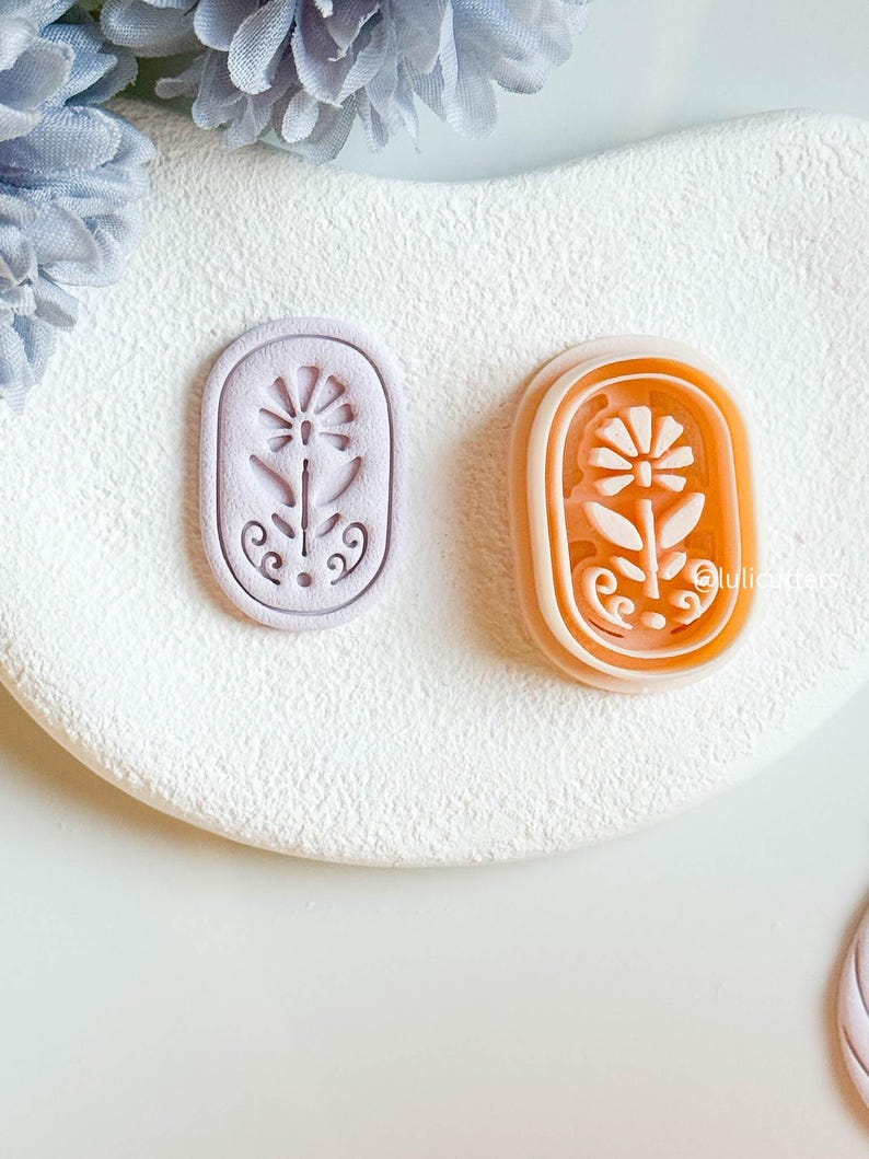 May include: Two floral-shaped cookie cutters. One is lavender, the other is orange with a white interior. Both are oval-shaped with a flower and stem design. The cutters are on a white, textured surface.