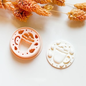 May include: A light pink cookie cutter with a coffee cup design, featuring a circular frame with cloud and dot cutouts. Next to it is a white cookie with the coffee cup design. Dried orange flowers are in the background.